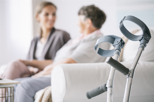 Unterarmgehstützen lehnen vor einem Sofa; im Hintergrund berät eine Pflegekraft eine ältere Frau während der Kurzzeitpflege.