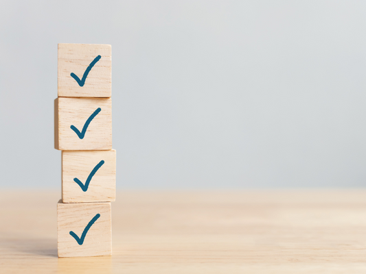 Vier Holzwürfel gestapelt auf einem Tisch, jeder Würfel zeigt ein blaues Häkchen, symbolisieren Erfüllung der Voraussetzungen für eine Pflegebox