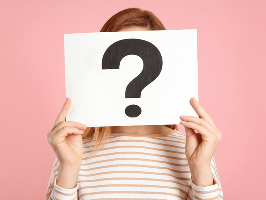 Person hält ein Schild mit einem schwarzen Fragezeichen vor ihrem Gesicht vor rosa Hintergrund; symbolisiert Fragen zum Pflegebox Anbieterwechsel